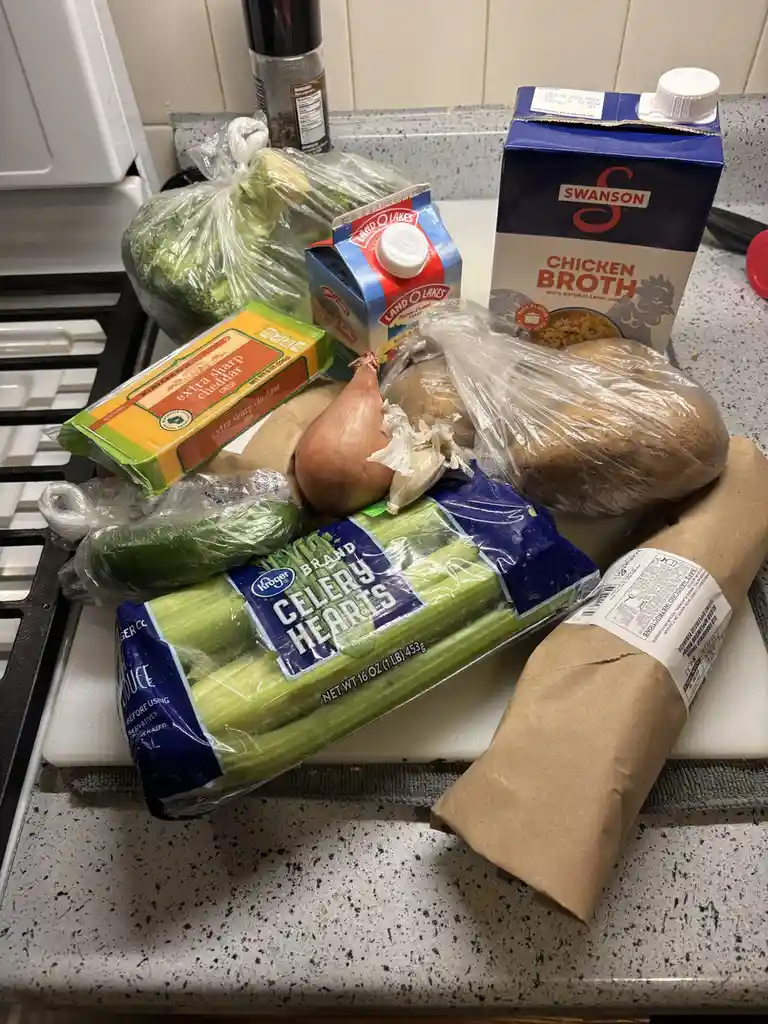 A kitchen counter displays a large assortment of groceries including celery hearts, cheddar cheese, chicken broth, potatoes…