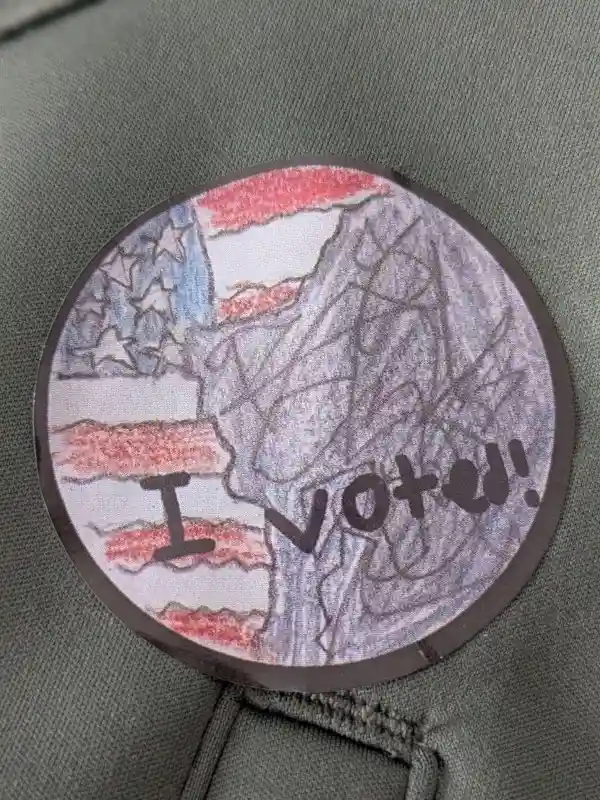 A close-up of a hand-drawn, childlike 'I Voted!' sticker on a green fabric surface, featuring a crude American flag design…