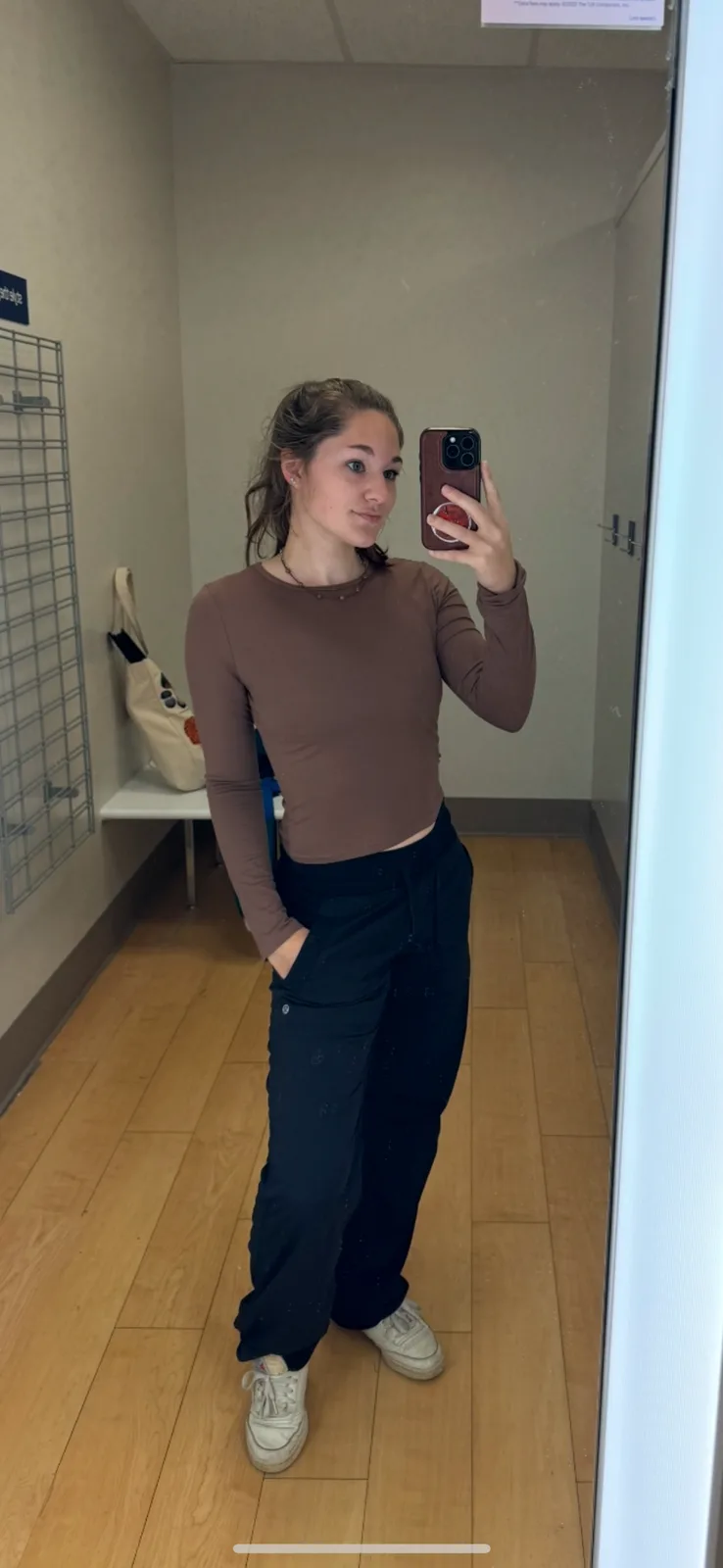 A young woman takes a mirror selfie in what appears to be a clothing store fitting room, wearing a brown long-sleeved crop…