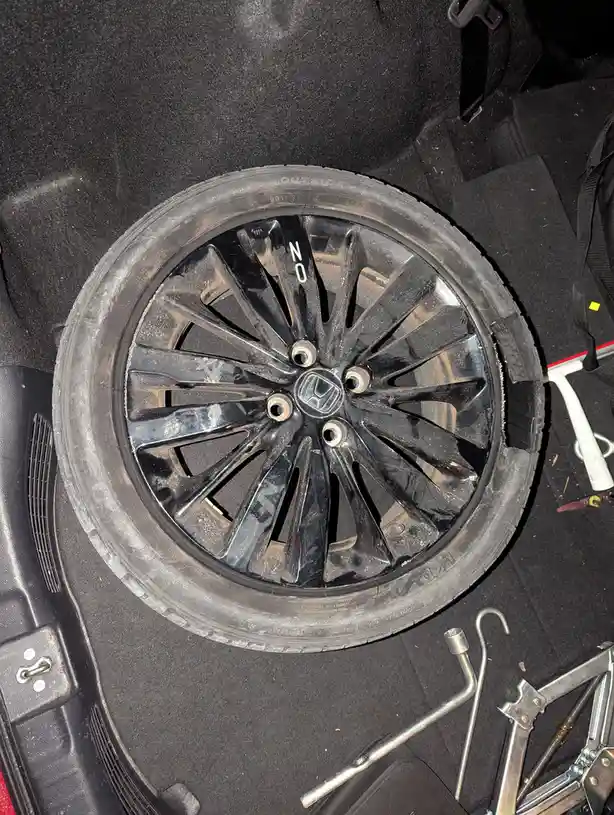 A dirty spare tire with a black Honda wheel rim is shown inside a car's trunk, surrounded by tools like a jack and lug…