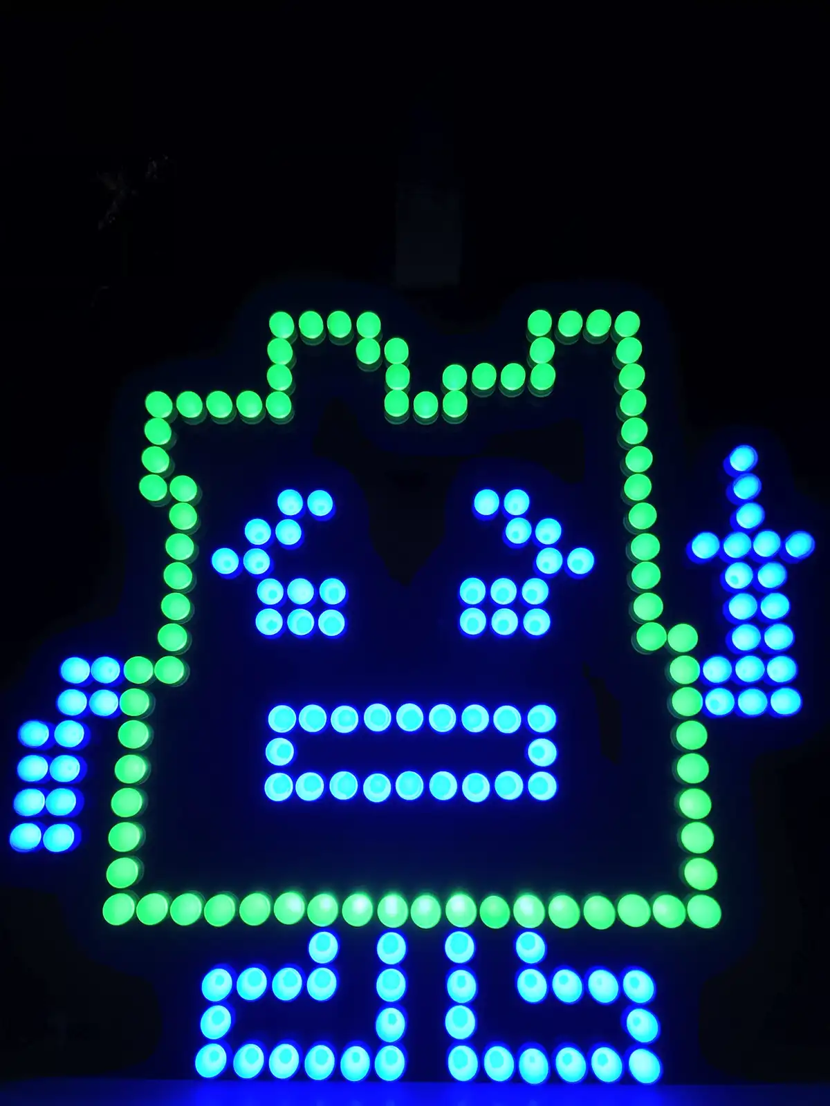 A pixelated LED sign shaped like a cartoon character with green outline and blue inner details, displaying the text 'mfw' at…
