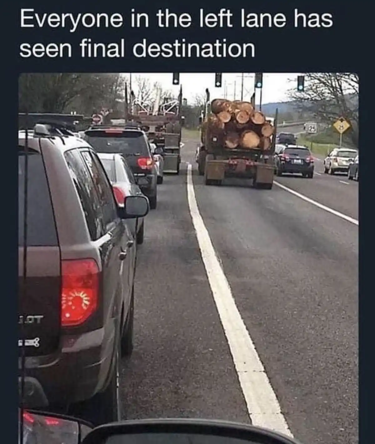 A photo from inside a car shows traffic on a multi-lane road, with cars stopped in the right lanes and a large log truck in…