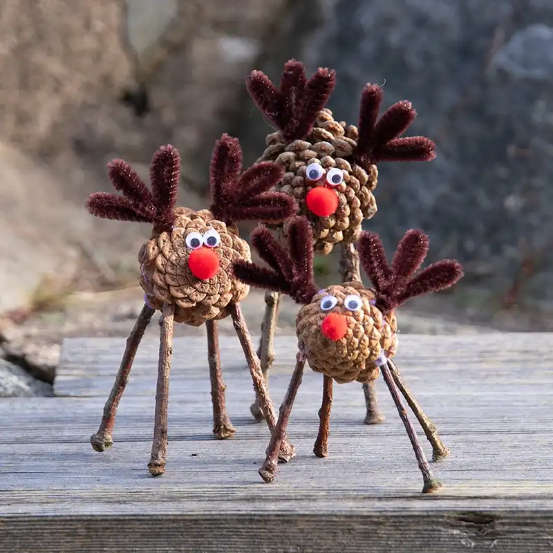 Three handmade reindeer crafts made from pinecones, pipe cleaner antlers, twig legs, googly eyes, and red pom-pom noses…