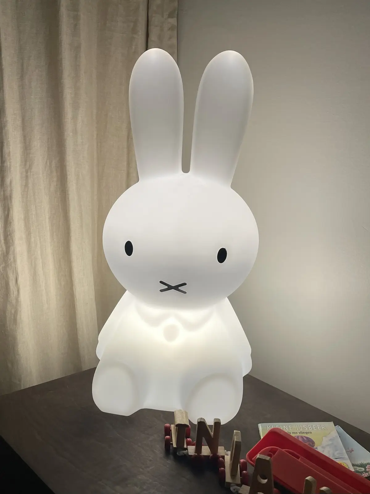 A white, glowing Miffy lamp shaped like a sitting rabbit with black dot eyes and an X-shaped mouth sits on a dark wooden…