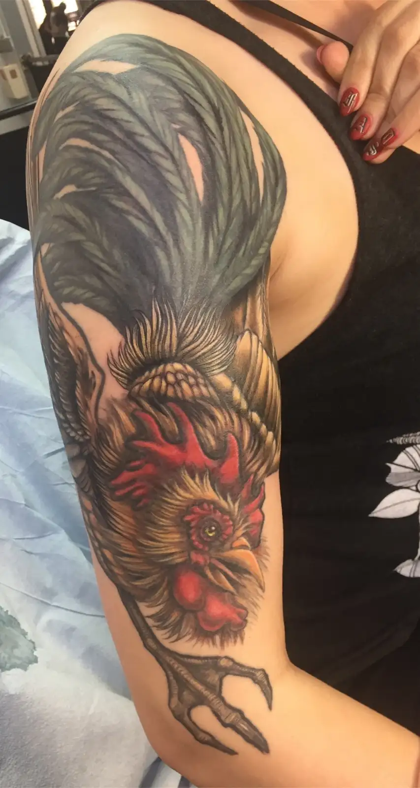 A person's left arm features a large, realistic tattoo of a rooster, with detailed feathers, a red comb, and a clawed foot…