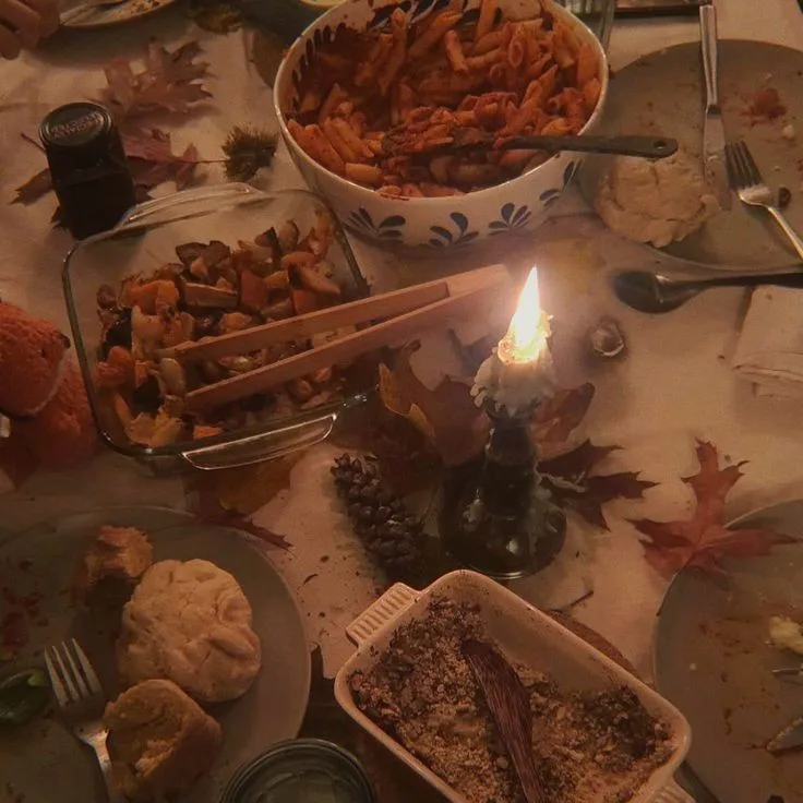 A dimly lit, messy dining table covered in autumn leaves, pinecones, and partially eaten dishes including pasta, roasted…