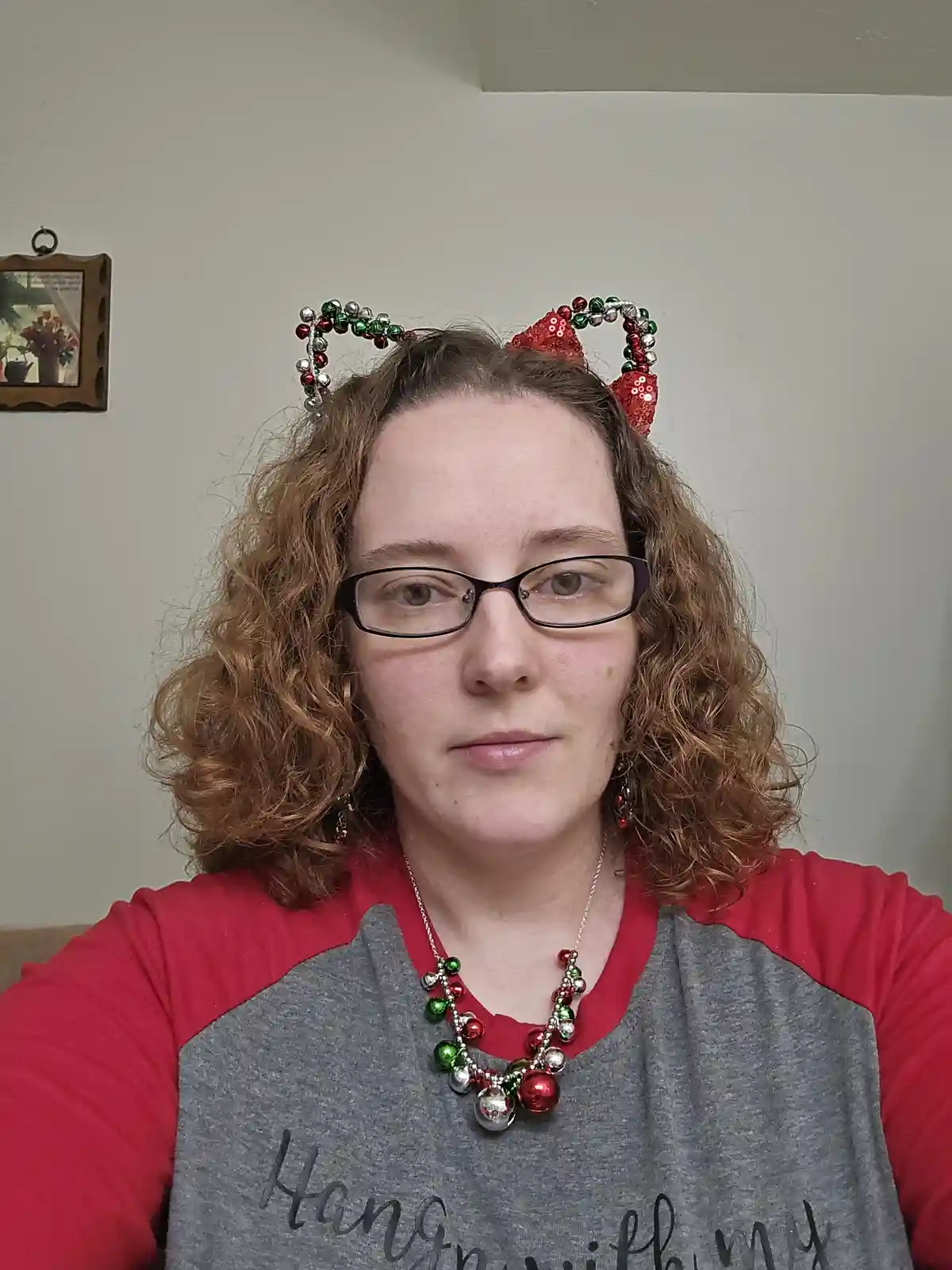 A person with curly brown hair and glasses wears a red and green holiday-themed headband with cat ears and a matching…