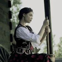 A woman in traditional folk attire sits on a wooden structure, holding a pole or stick. She is shown in six sequential…