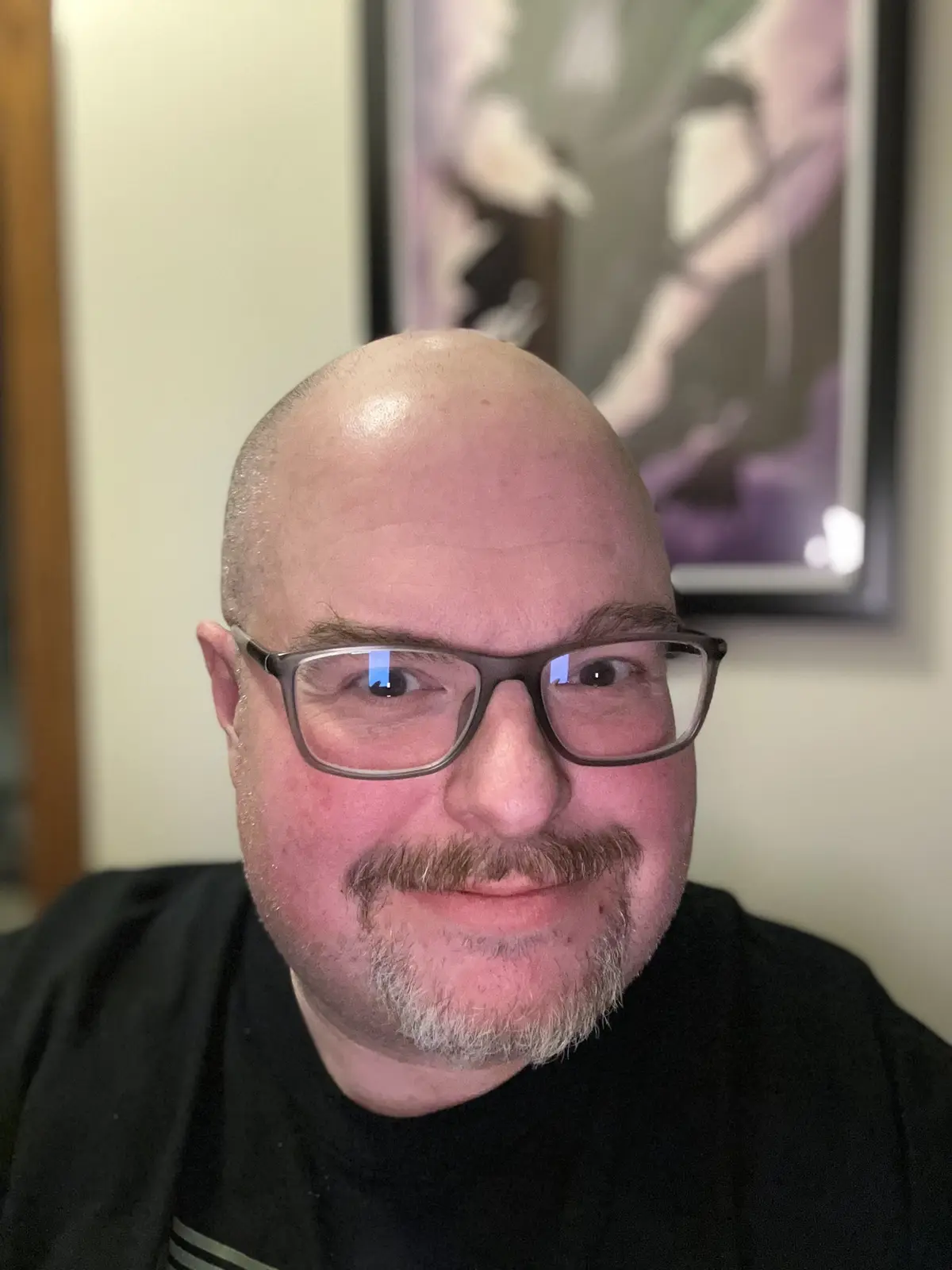 A close-up photo of a bald man with glasses and a goatee, wearing a black shirt, looking directly at the camera with a…
