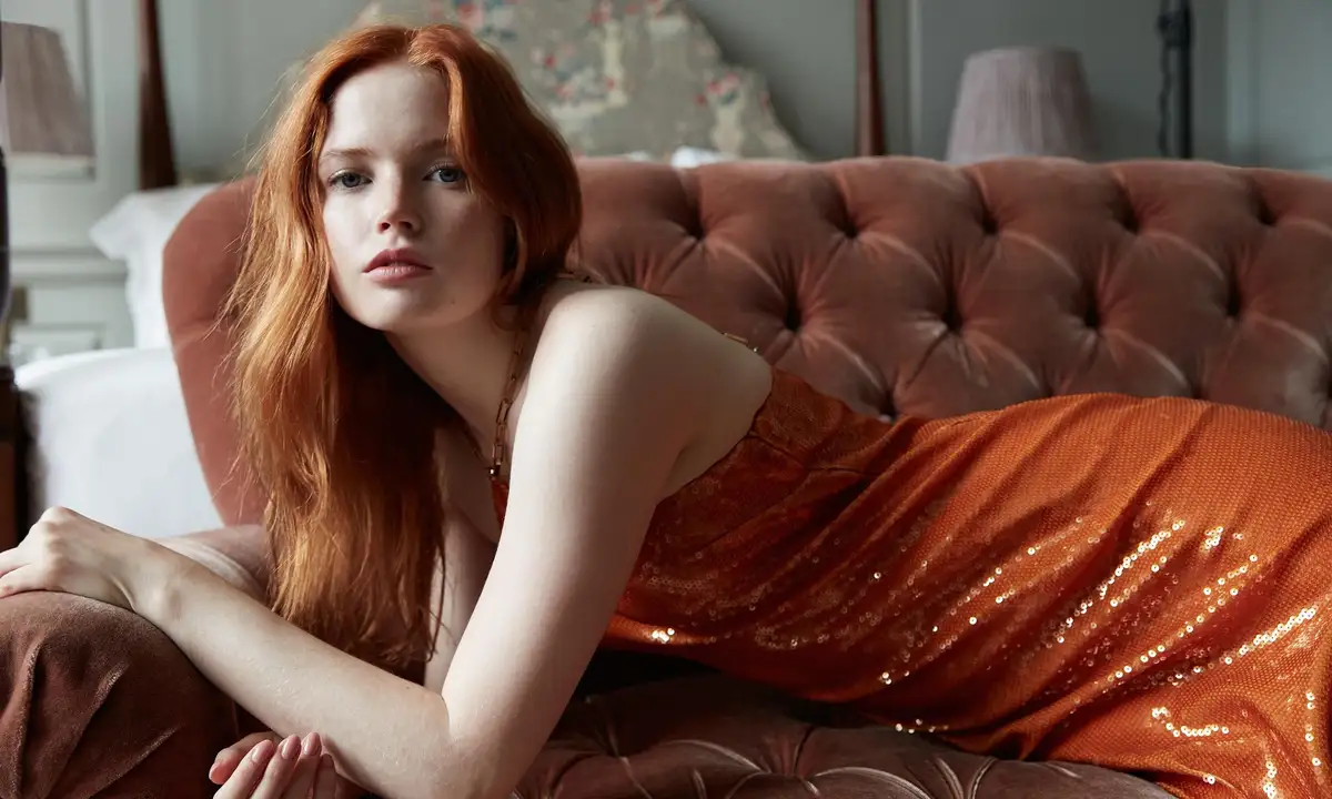 a woman with red hair wearing an orange sequined dress lies on a brown tufted velvet couch in a bedroom. She gazes directly…