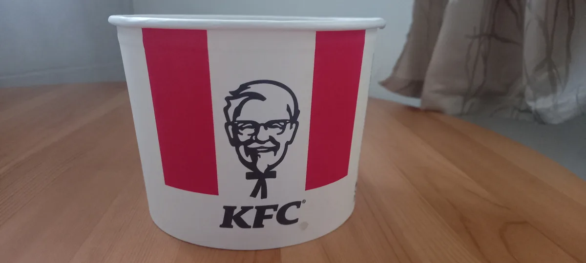 A classic KFC chicken bucket with the Colonel Sanders logo and red stripes sits on a wooden table. The bucket is empty and…