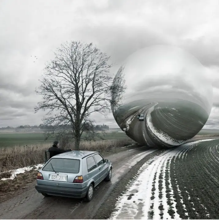 A silver car is parked on a snowy rural road next to a bare tree, facing a giant, reflective metallic sphere that appears to…