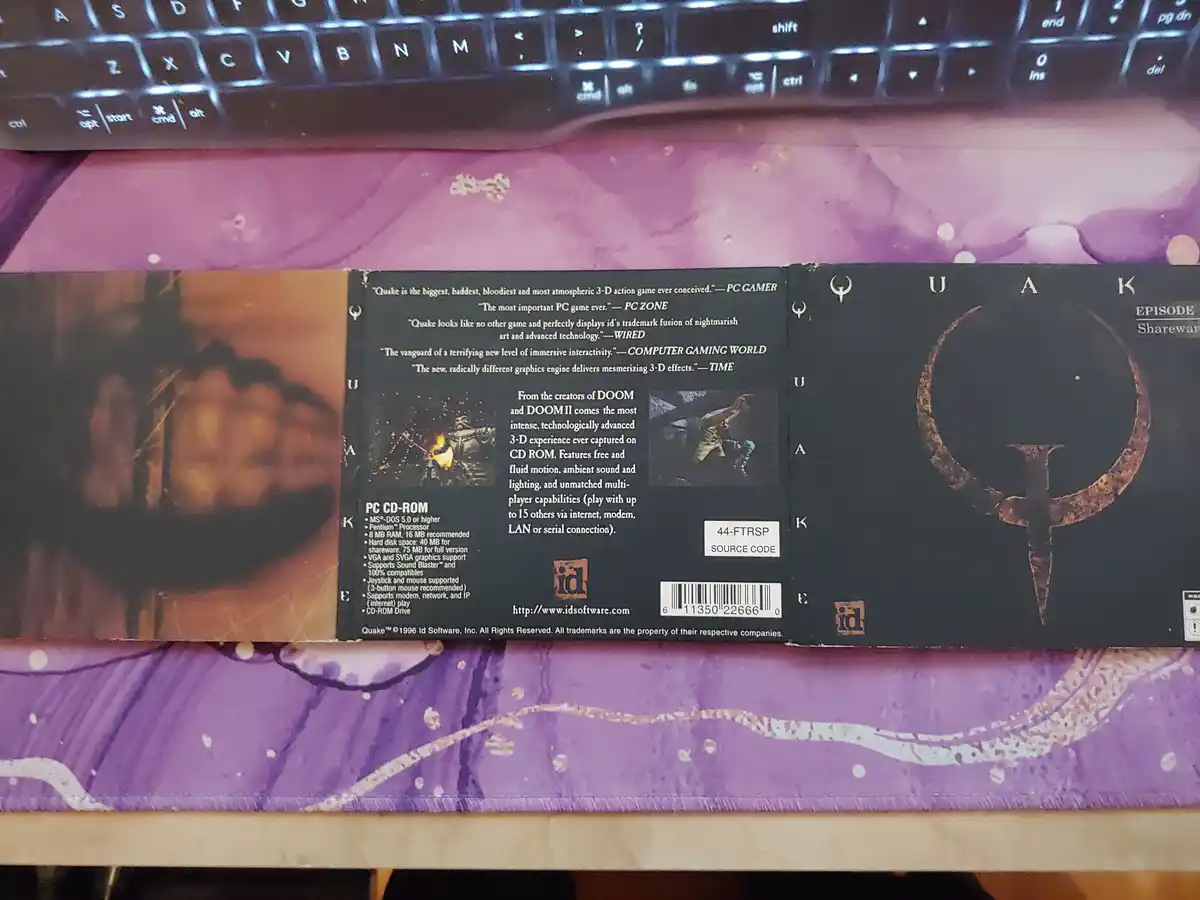 The back of a Quake Episode Shareware PC CD-ROM game case is shown lying on a purple desk mat, with a black keyboard visible…