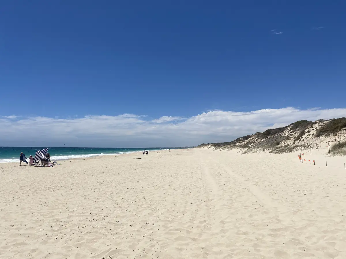A wide, sunny beach with light-colored sand, turquoise ocean, and a clear blue sky with scattered clouds. A few people are…