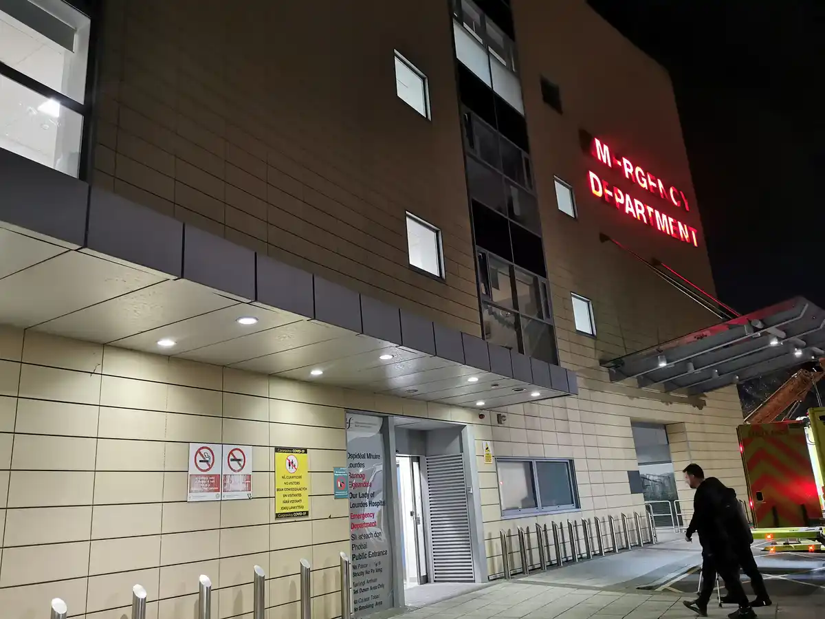 A nighttime photo of a hospital's emergency department entrance, where a red neon sign above the building reads 'M-RGNCY…