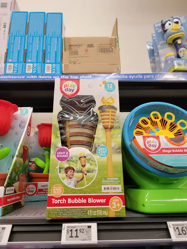 A product shot of a 'Play Day' Torch Bubble Blower toy in its packaging, displayed on a store shelf next to other bubble…