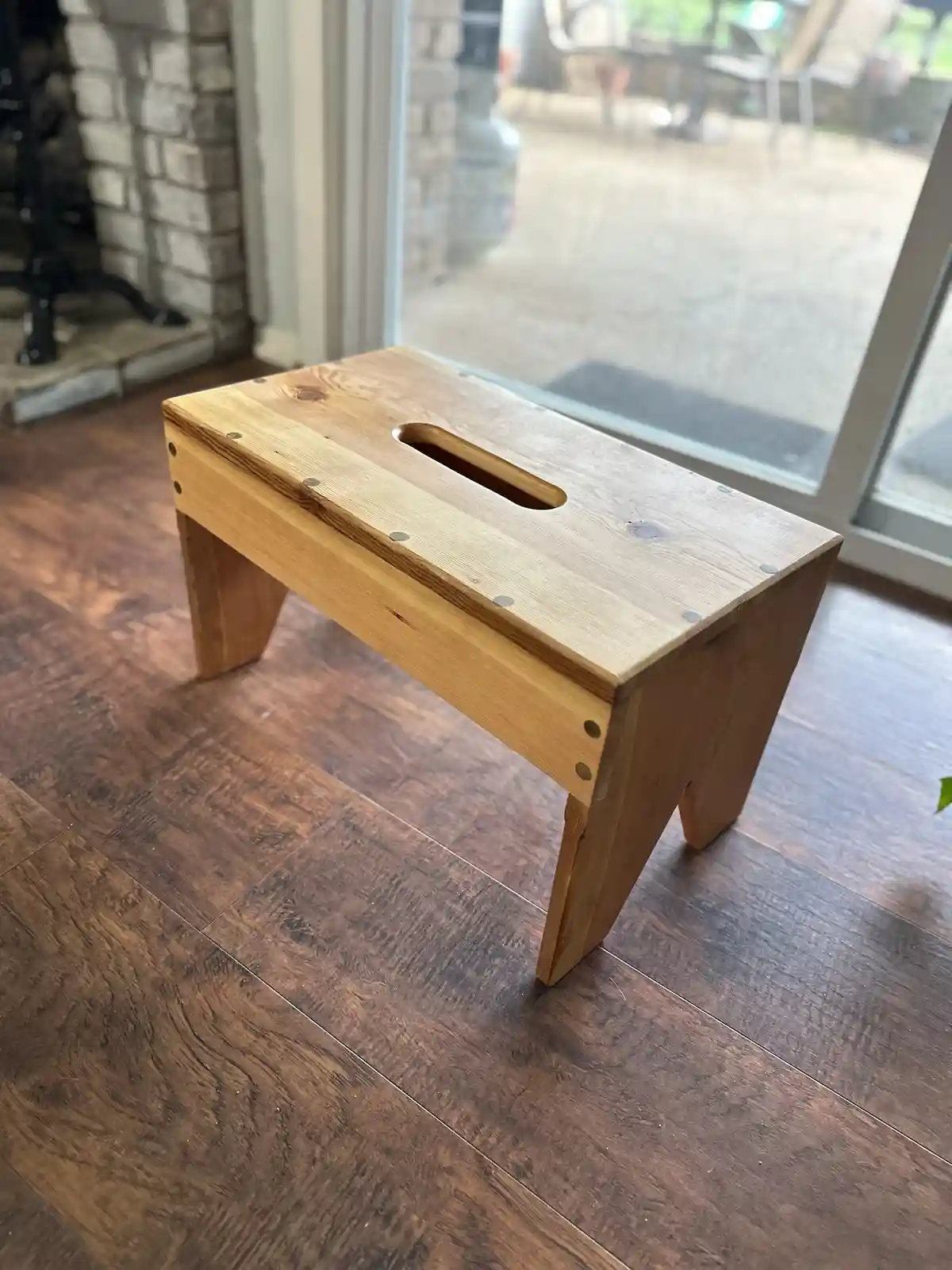 A simple wooden stool with a rectangular top and four legs sits on a dark wood floor near a glass door. The top of the stool…