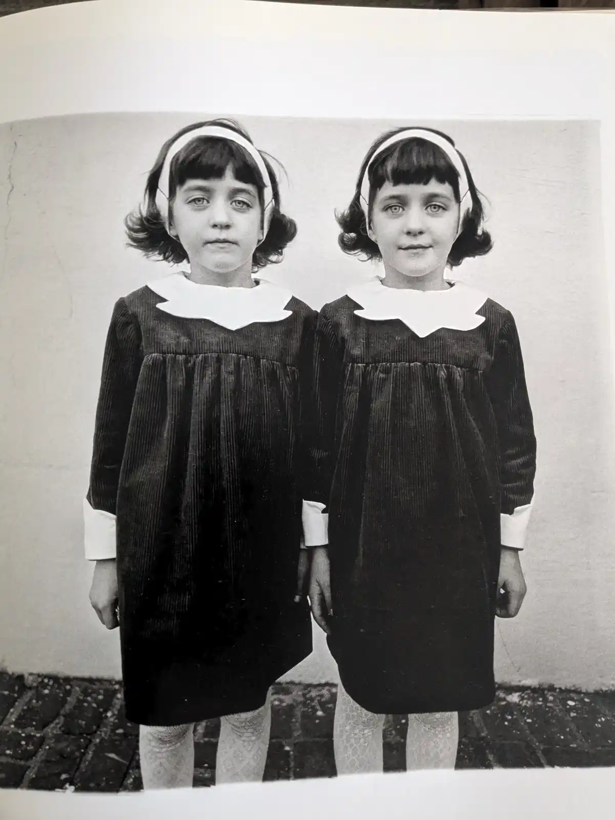 A vintage black-and-white photograph shows two young girls standing side-by-side, dressed identically in dark corduroy…