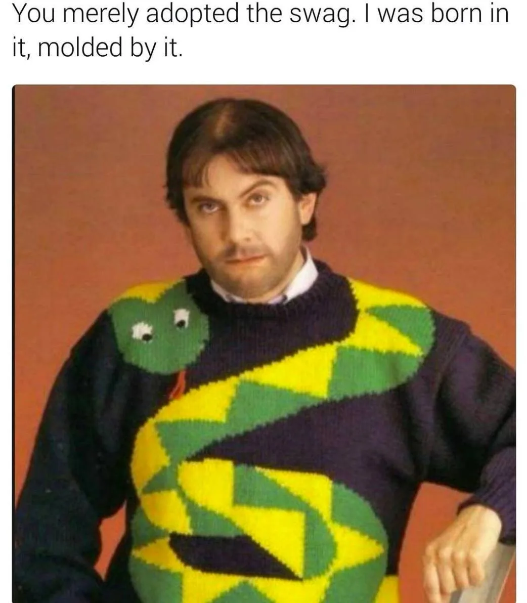 A man with dark hair and a mustache stares intensely at the camera while wearing a dark blue sweater with a large, colorful…