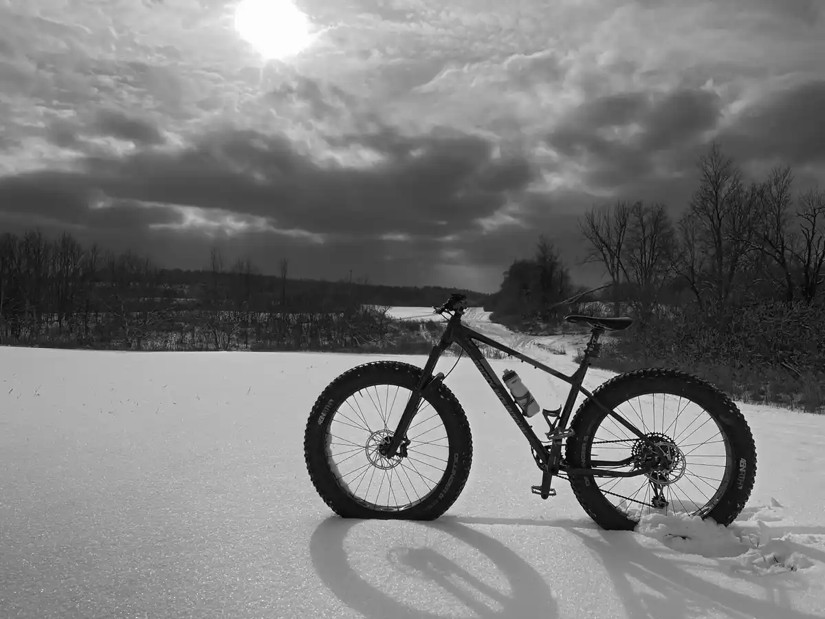 A black-and-white photo shows a fatbike with wide tires standing upright in a snowy field under a cloudy sky with the sun…