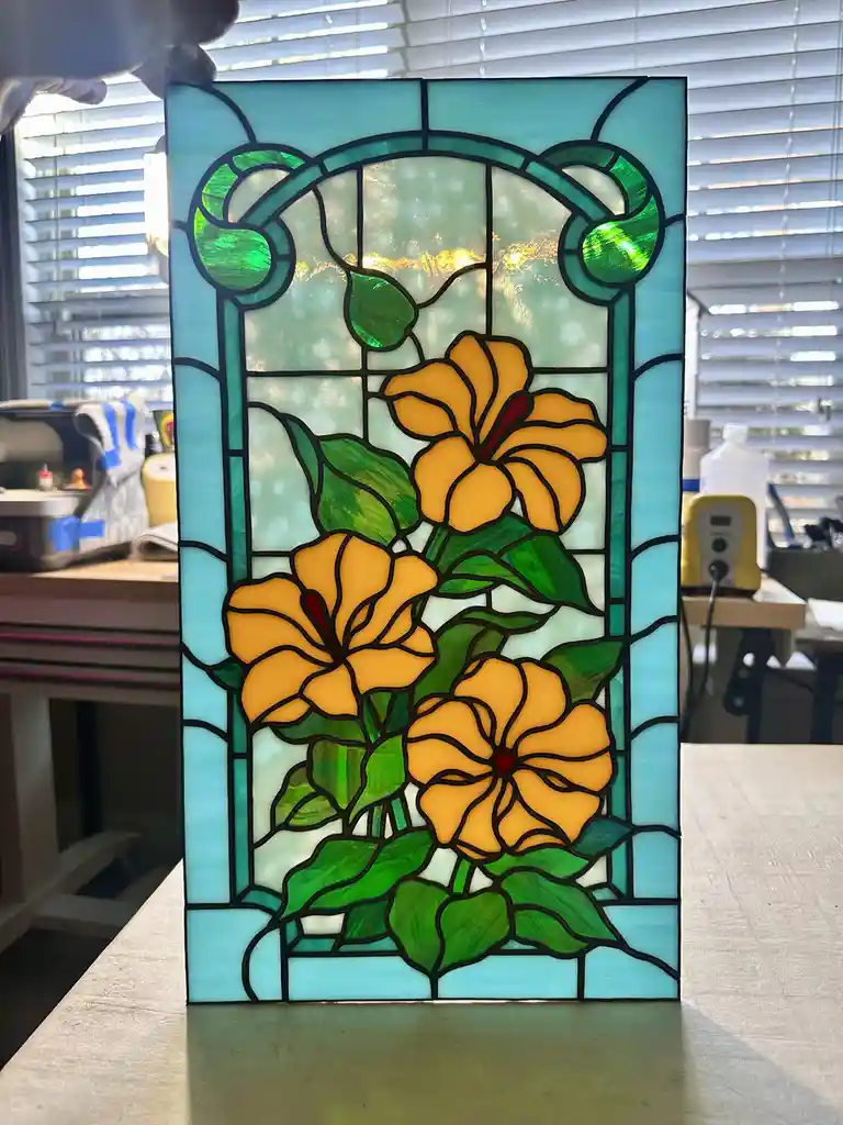 A handmade stained glass panel featuring three yellow flowers with green leaves and a teal border, standing upright on a…