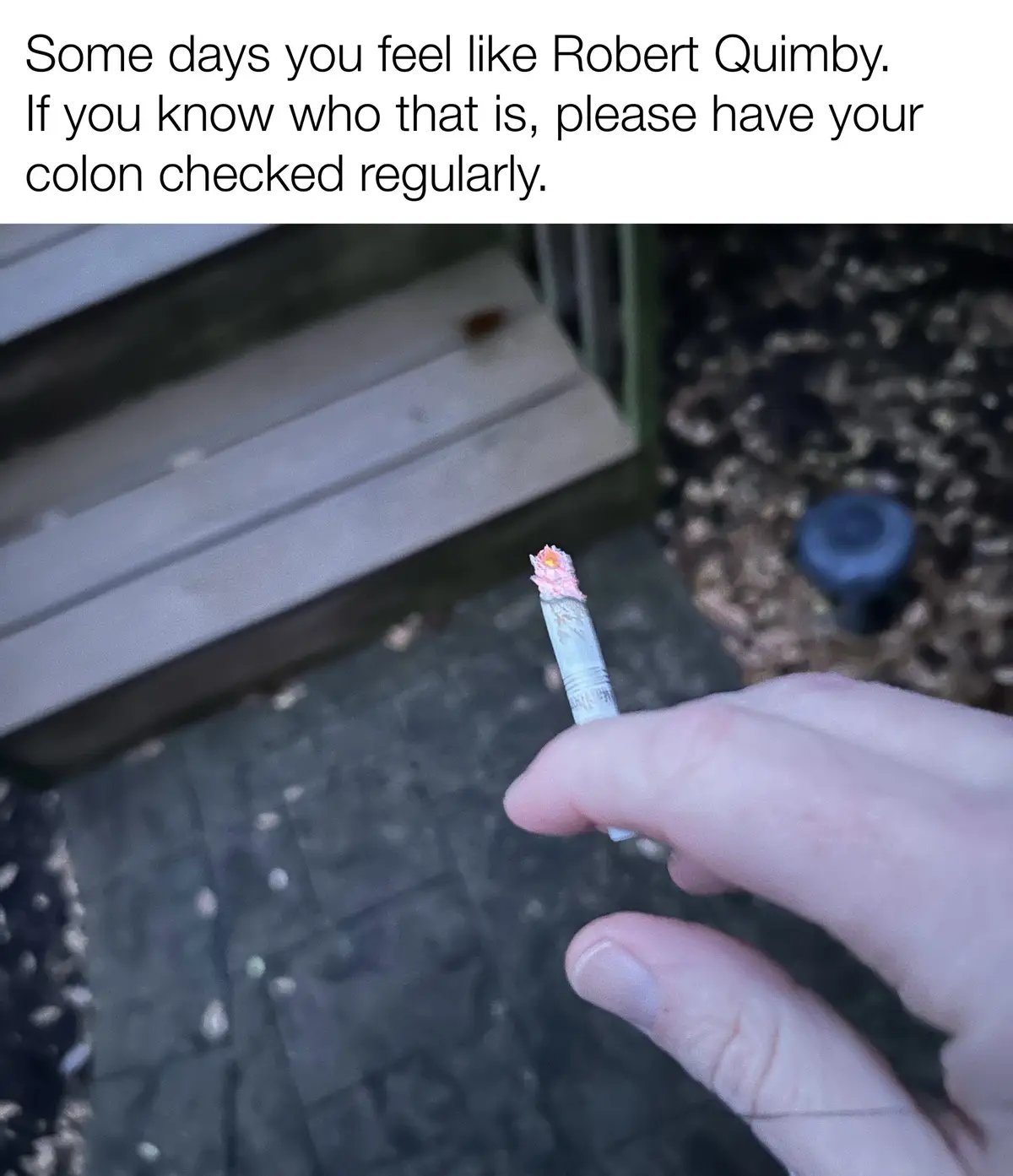 A close-up photo shows a hand holding a lit cigarette outdoors, with a blurred background of pavement and mulch. The…