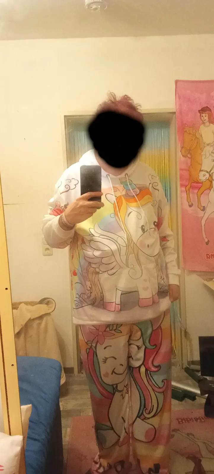 A person with their face obscured takes a mirror selfie wearing a full-body unicorn-themed pajama set, featuring a winged…