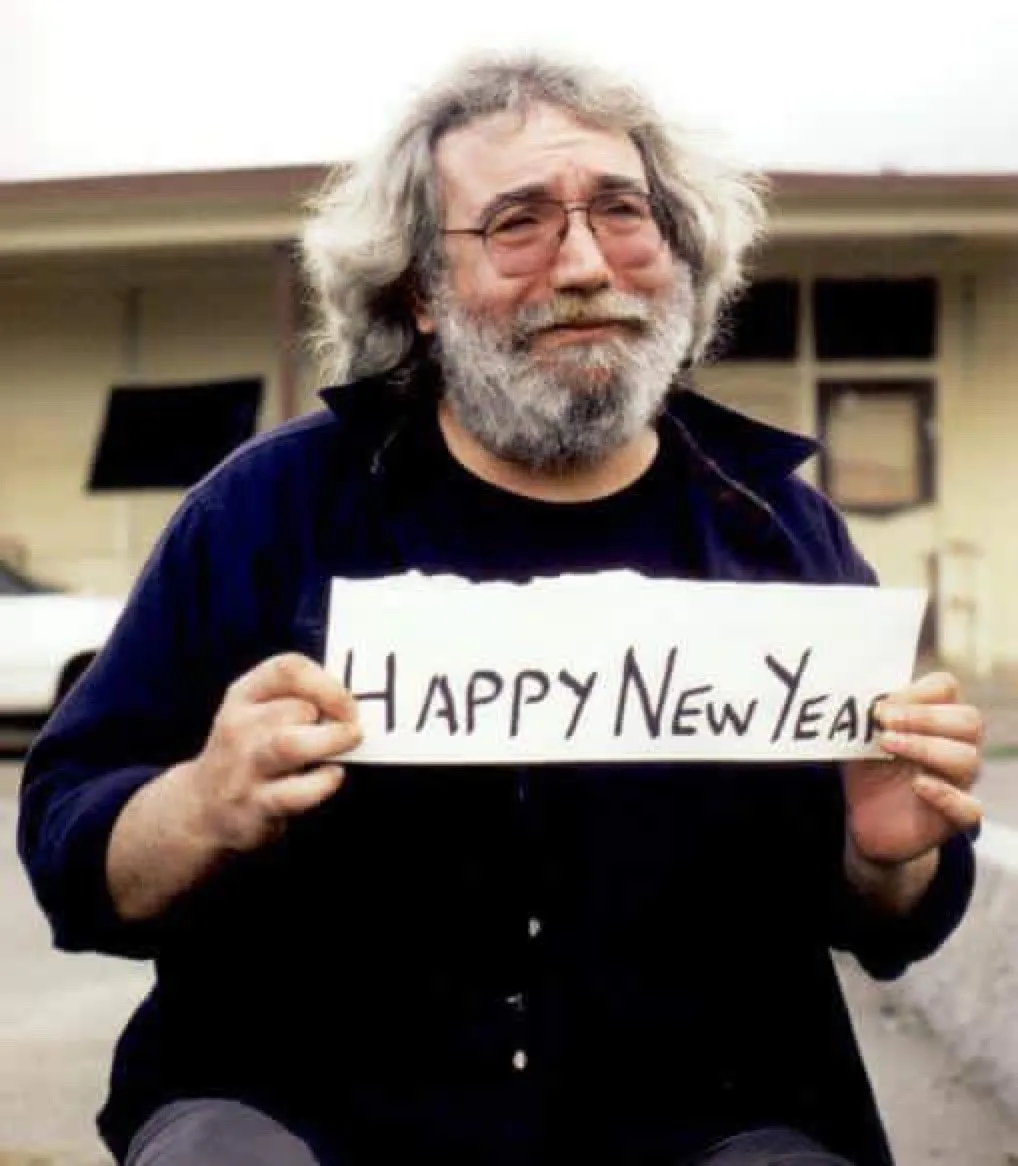 A man with long gray hair, glasses, and a beard holds a handwritten sign that reads 'HAPPY NEW YEAR' while looking directly…