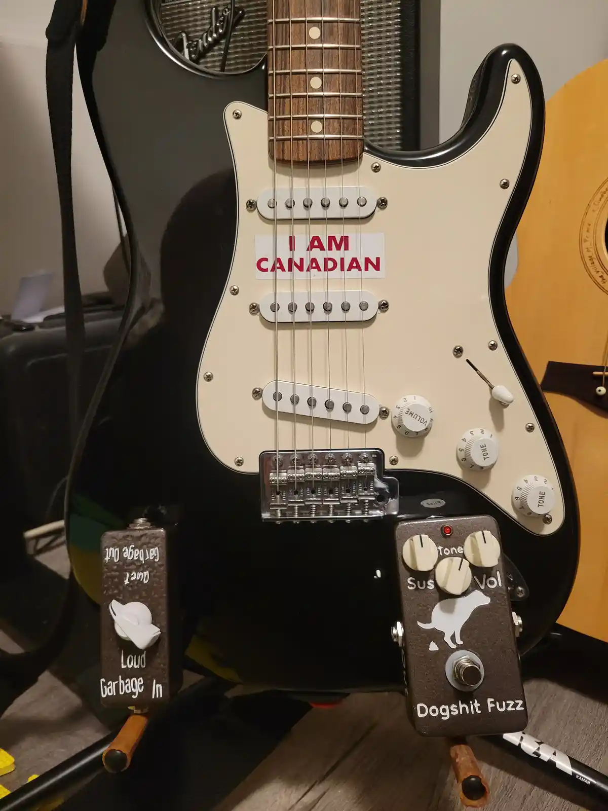 A black and cream Fender Stratocaster electric guitar has a sticker that reads 'I AM CANADIAN' on its body. Two guitar…