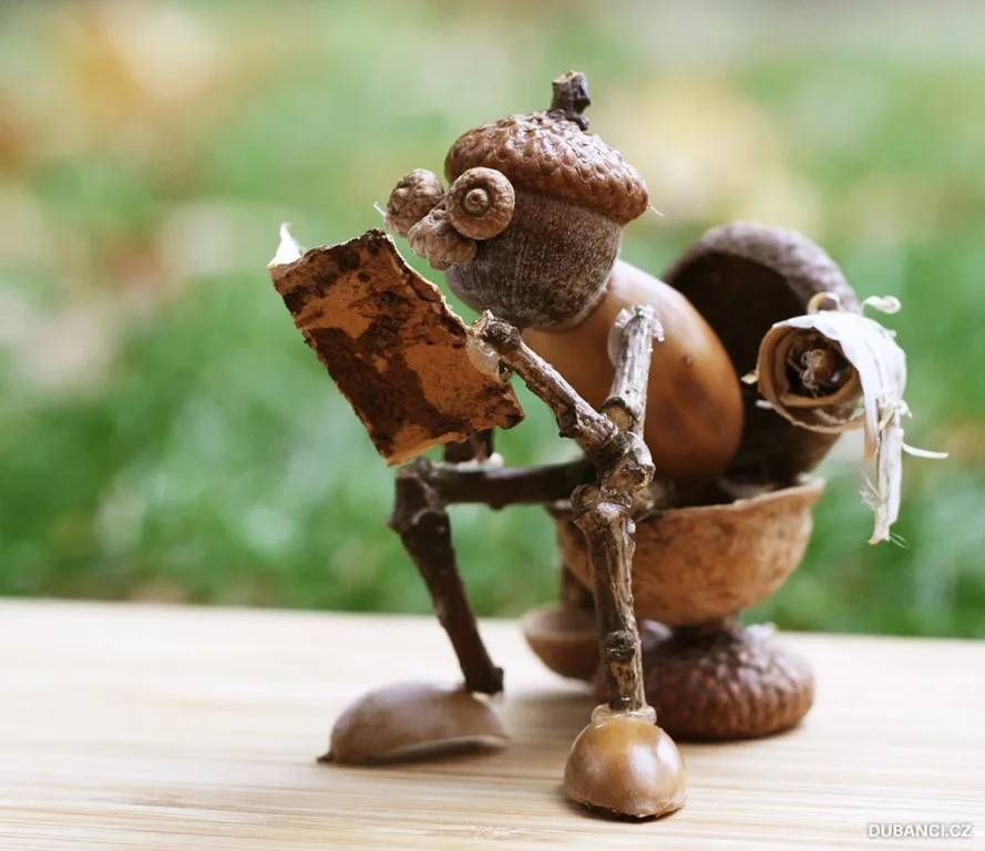 A whimsical sculpture made of acorns and twigs depicts a tiny figure sitting on a wooden surface, holding a small, weathered…