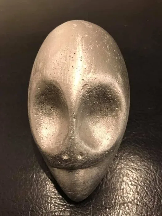 A close-up of a metallic, silver-colored alien head sculpture with large, dark eye sockets and a pointed chin, resting on a…