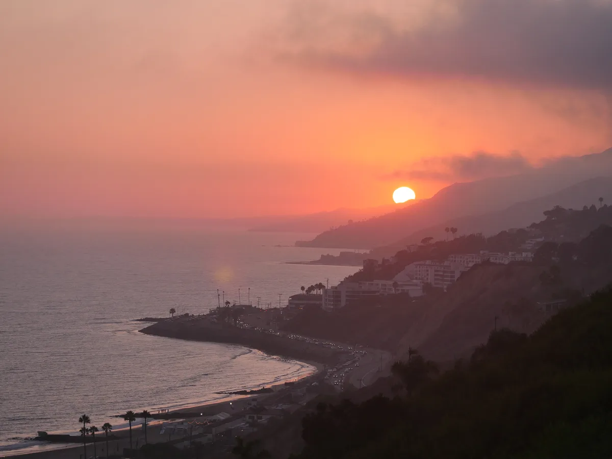 A scenic coastal sunset with the sun setting behind hills, casting orange and pink hues across the sky and reflecting on the…