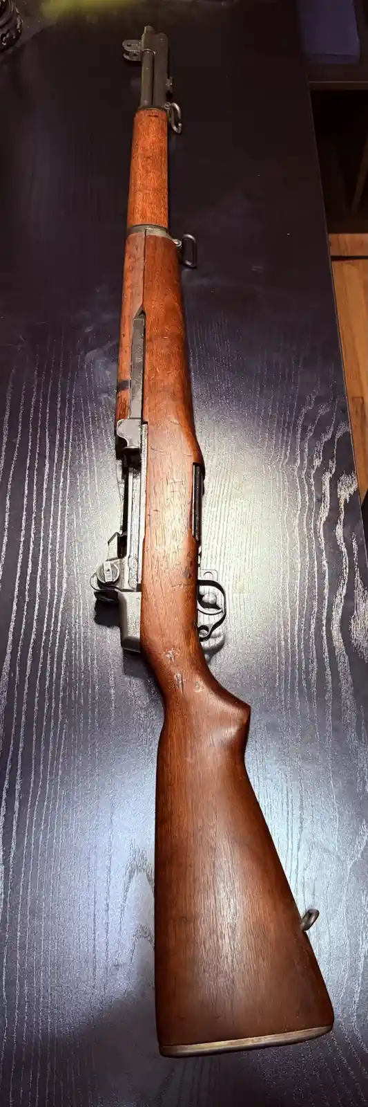 A full-length M1 Garand rifle with a wooden stock and metal components lies horizontally on a dark wood-grain table. The…