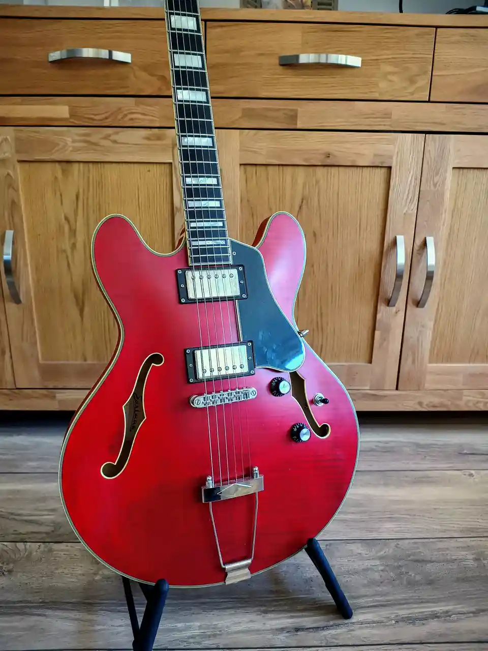 A red semi-hollow body electric guitar with f-holes and a black pickguard stands upright on a black stand against a wooden…
