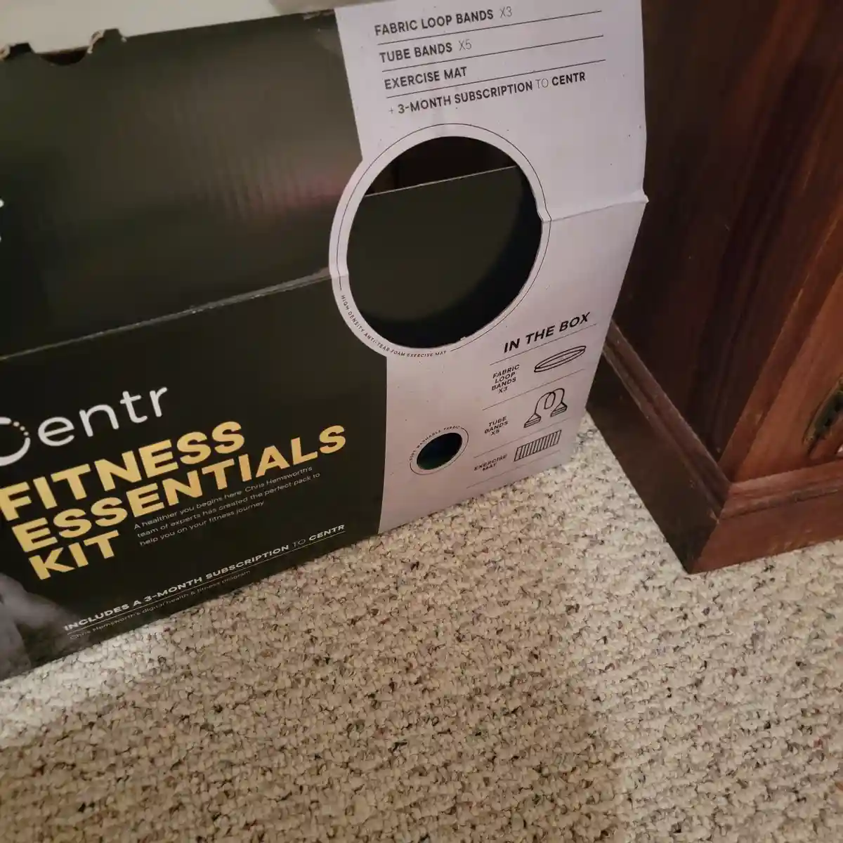 A close-up photo of a Centr Fitness Essentials Kit box sitting on a carpeted floor next to a wooden cabinet. The box lists…
