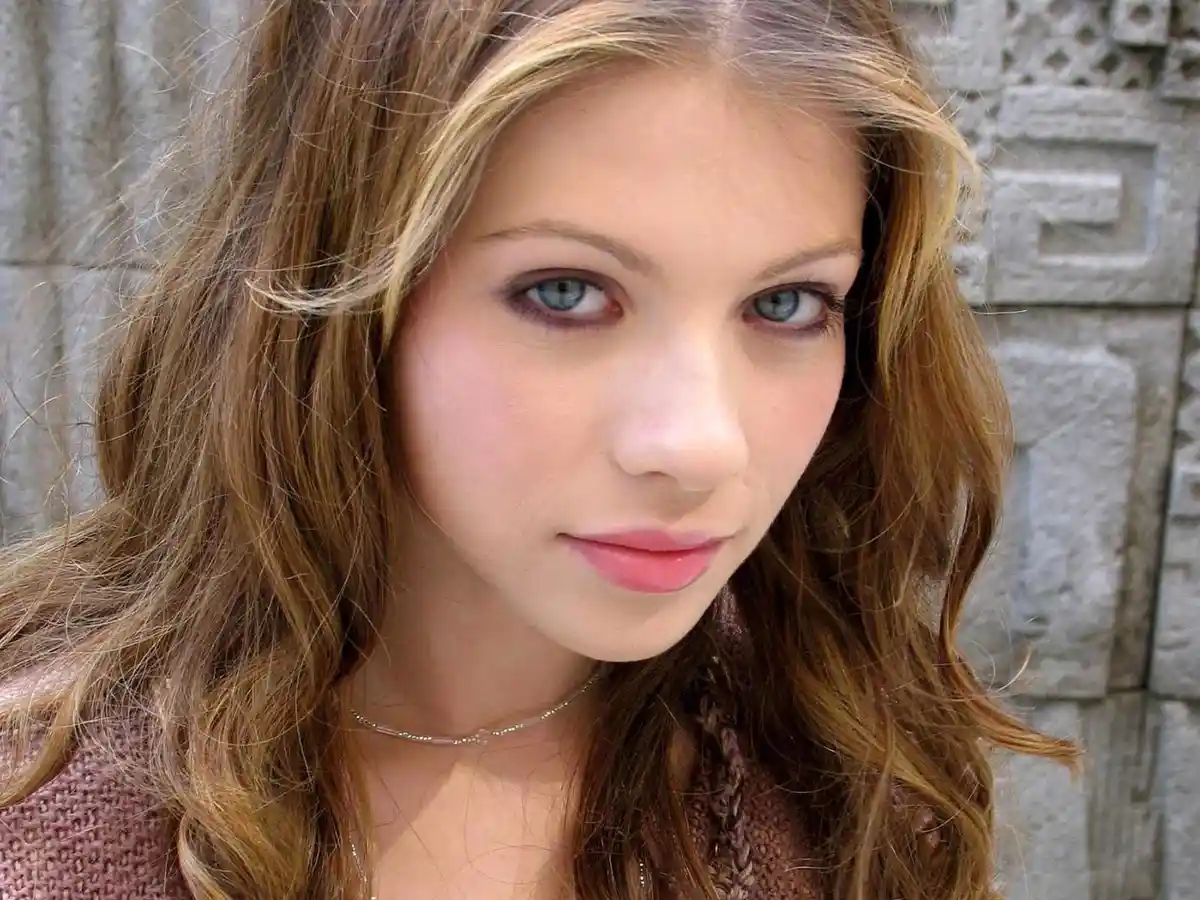 A close-up photo of a young woman with long, wavy brown hair and blue eyes, wearing a pink sweater and a thin necklace…