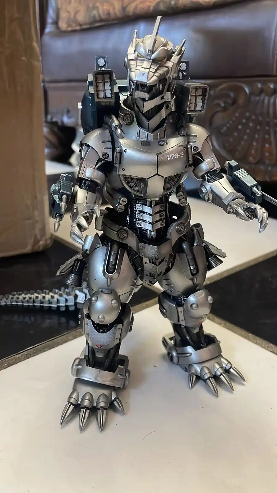 A highly detailed, silver and black armored Godzilla action figure stands on a white surface, its chest plate clearly marked…