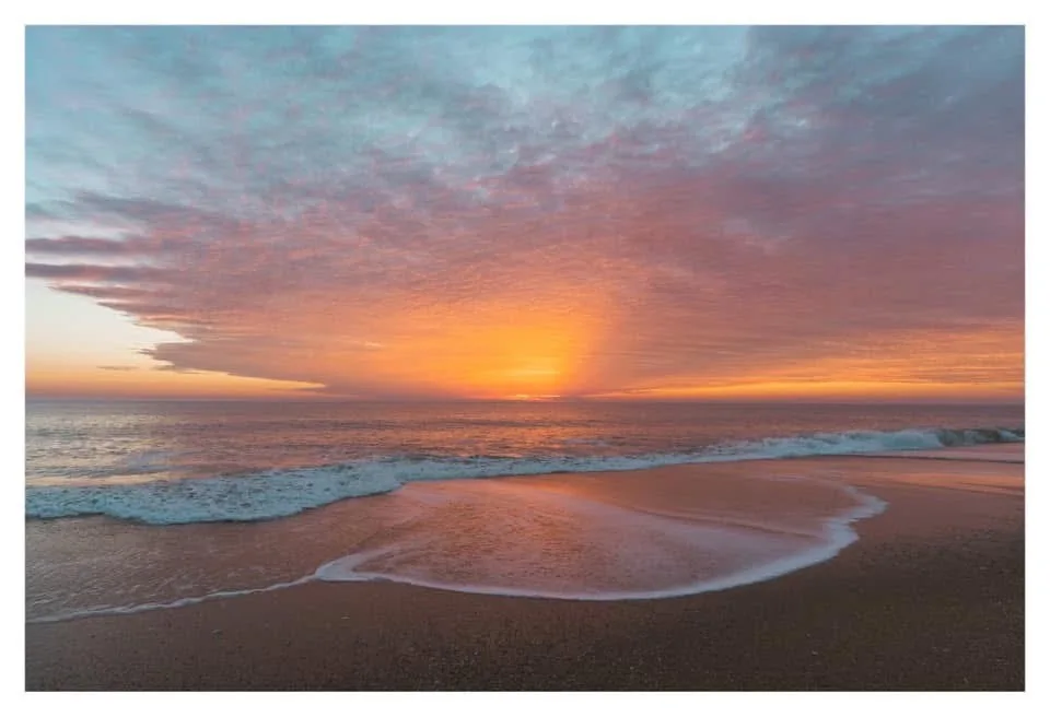 A serene beach sunset with orange and pink clouds over the ocean, gentle waves washing onto sandy shore. Overlaid text reads…