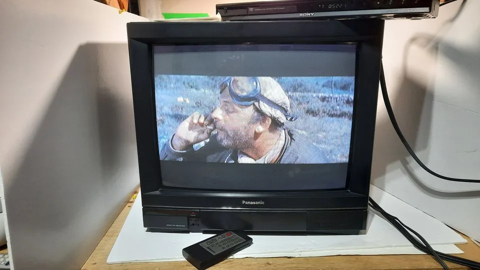 A vintage Panasonic CRT television displays a scene from a movie featuring a man with goggles smoking, while a Sony VCR sits…