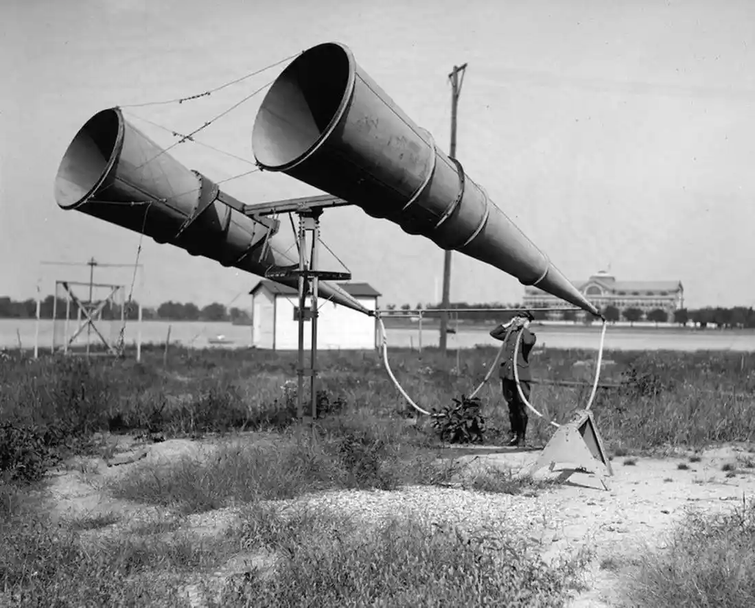 A black-and-white historical photo shows a man standing in a grassy field, holding a hose connected to a large, dual-cone…