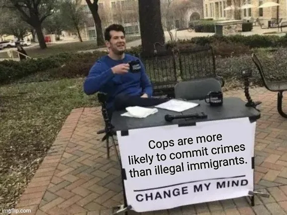 A man in a blue sweatshirt sits at a small outdoor table with a sign that reads 'Cops are more likely to commit crimes than…