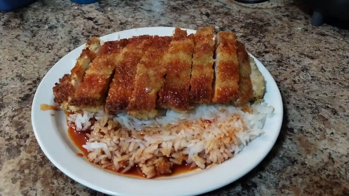 A white plate holds a serving of sliced, golden-brown breaded katsu resting on a bed of white rice mixed with red sauce…