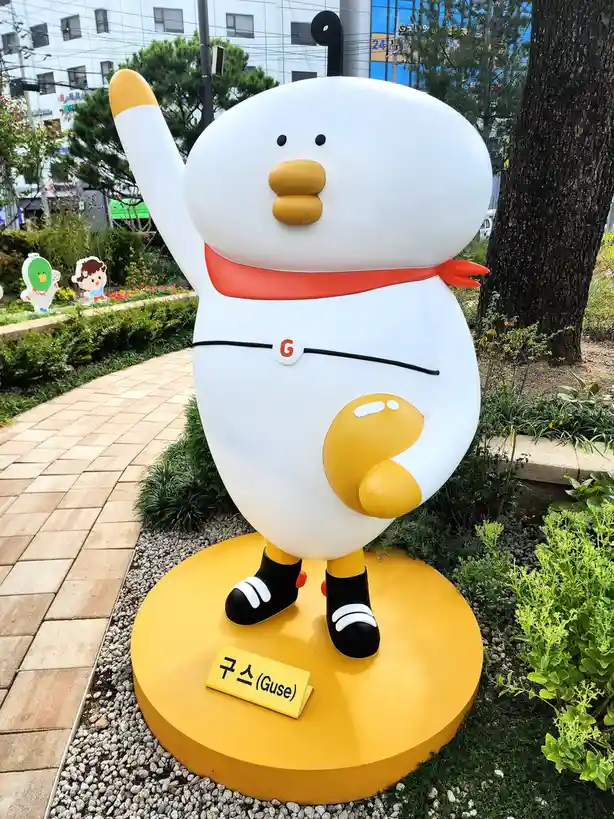 A large outdoor statue of a white, egg-shaped cartoon character named Guse, waving with one arm and wearing a red scarf and…
