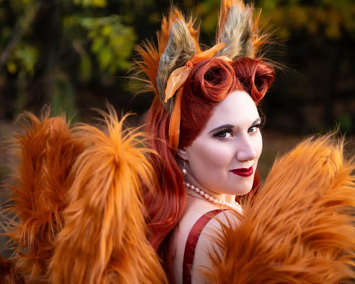 A woman with red hair styled in curls wears orange and grey fox ears tied with an orange ribbon and a large, fluffy orange…