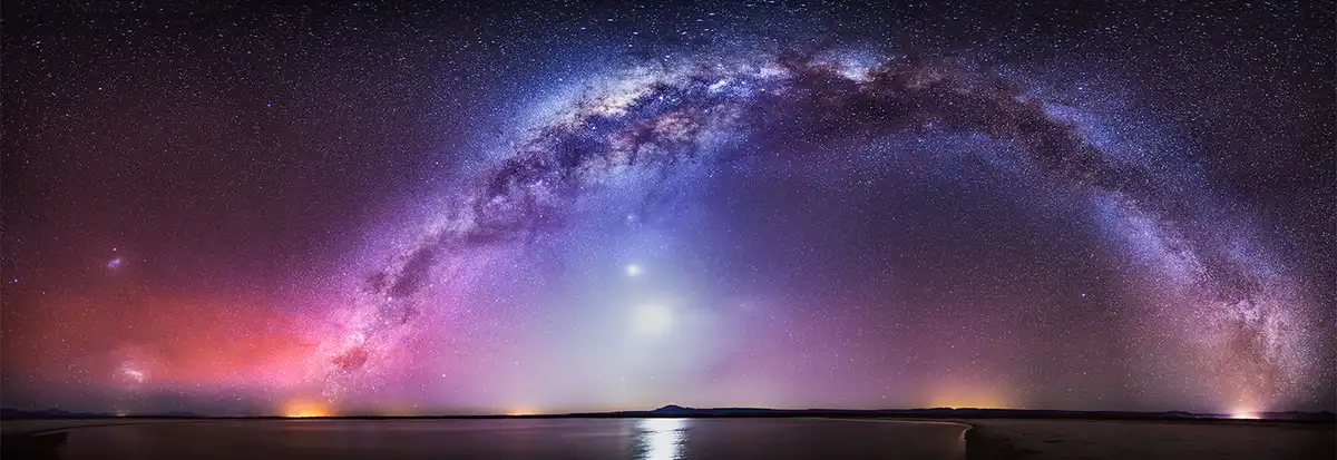 A panoramic night sky photograph shows the Milky Way galaxy arching across a star-filled purple and pink sky over a calm…