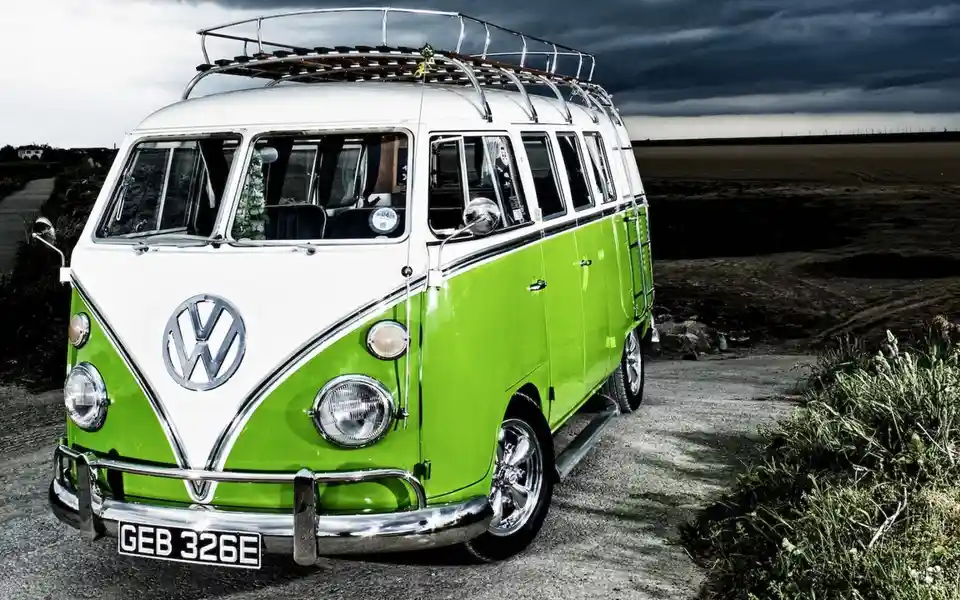 A vintage white-and-lime-green Volkswagen Type 2 van with a chrome roof rack is parked on a gravel road under a dark, stormy…