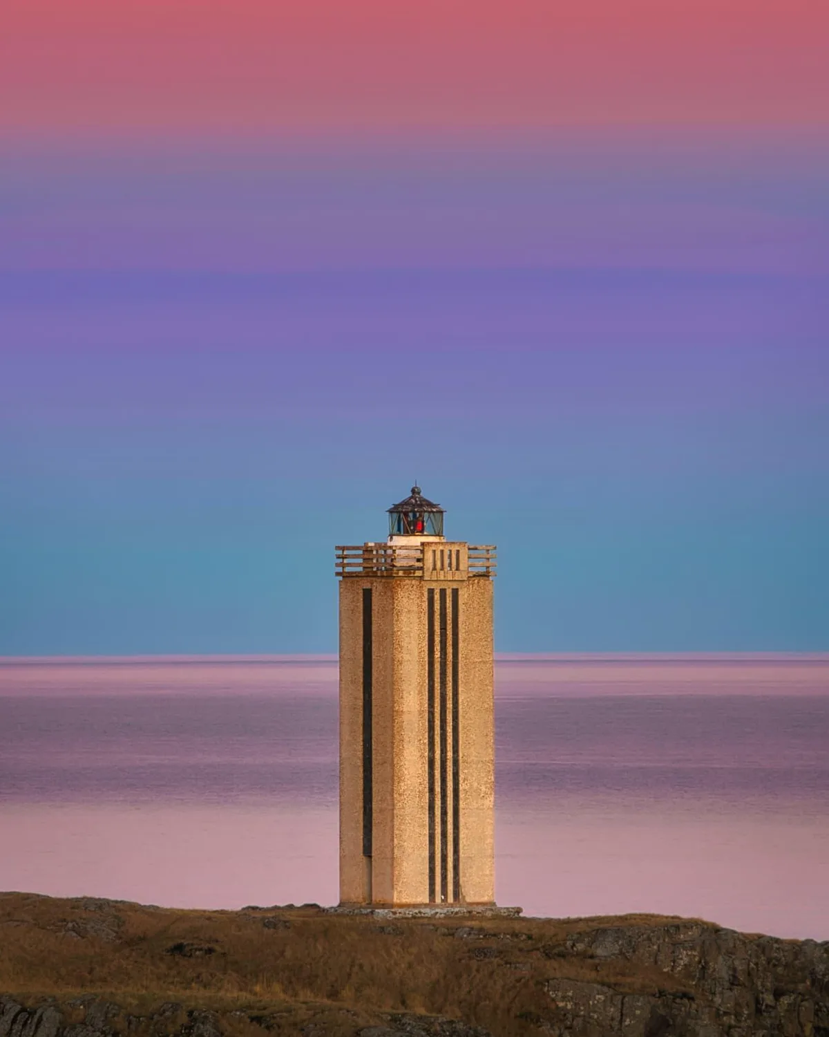 A tall, beige lighthouse stands on a rocky cliff overlooking a calm sea under a vibrant, gradient sky transitioning from…