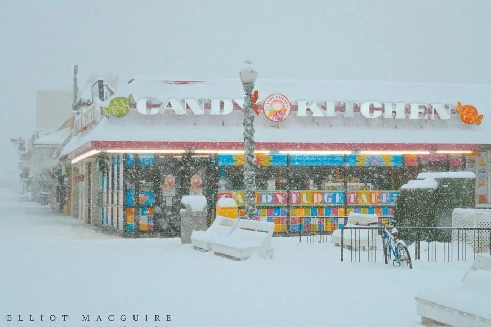 A snowy exterior shot of a brightly colored 'Candy Kitchen' storefront, its roof and surrounding benches covered in snow…