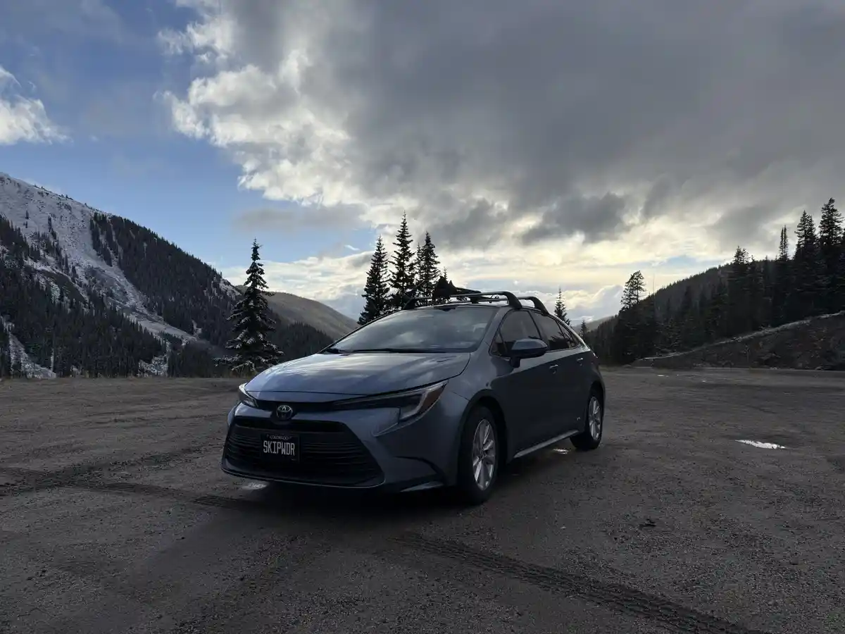 A gray Toyota Corolla parked in a snowy mountain parking lot with pine trees and cloudy skies. The Colorado license plate…