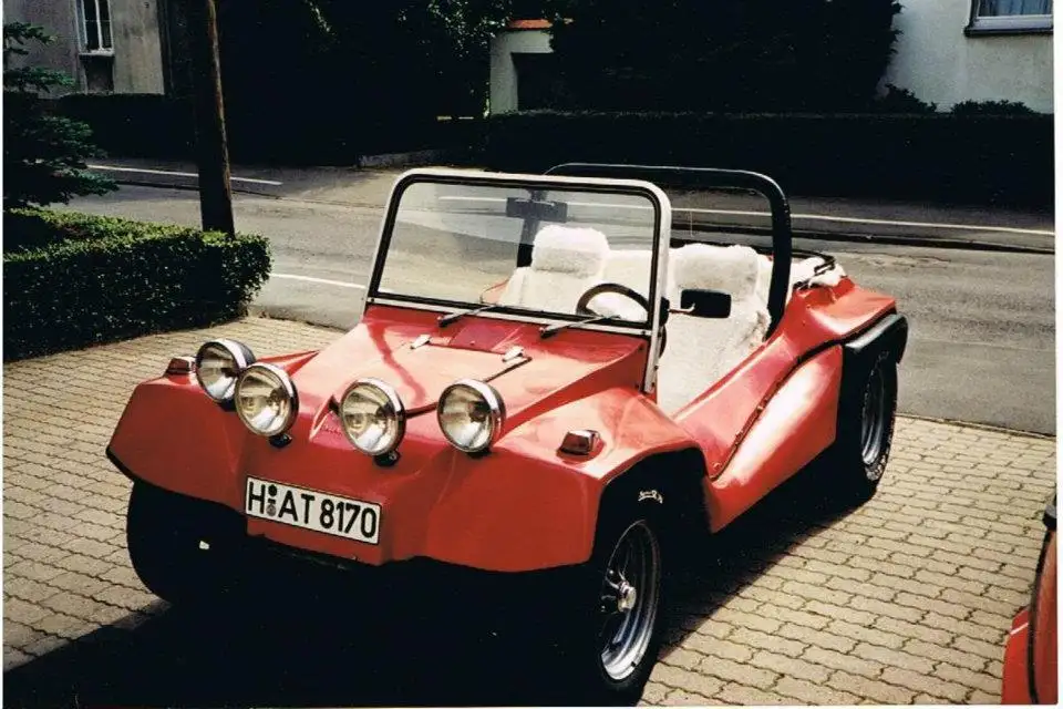 A red, open-top, minimalist dune buggy-style car with four round headlights and white seats is parked on a paved brick…